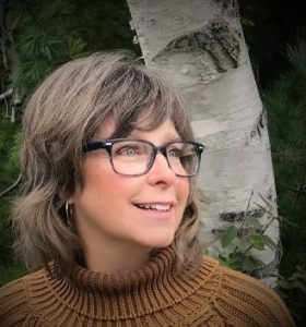 UPPAA A woman wearing glasses and a turtleneck sweater in front of a birch tree.