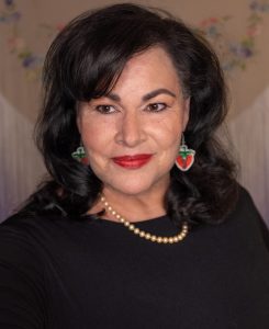 UPPAA A woman with shoulder-length dark hair and red lipstick is smiling at the camera, wearing a black top, pearl necklace, and red and green earrings. she stands against a background with floral patterns.