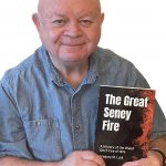 UPPAA A man holding a book titled "the great seney fire" while smiling at the camera.