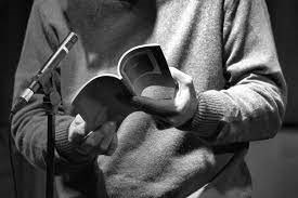 UPPAA A person in a sweater stands in front of a microphone, holding an open book in their hands.