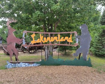 UPPAA A colorful outdoor sign for "lakemonoland" featuring stylized metal cutouts of a deer and a bear on either side of an orange, cursive script. the sign is set against a backdrop of trees.