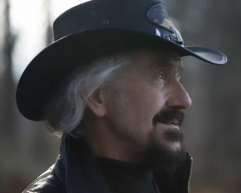 UPPAA A man with a gray beard and mustache wearing a black cowboy hat and leather jacket looks to the side. The background is blurred, suggesting an outdoor setting.
