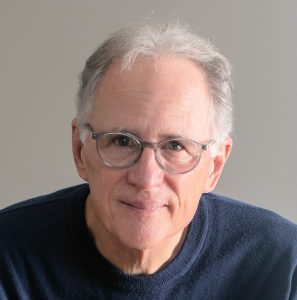 UPPAA An older man with short gray hair and glasses is wearing a navy blue sweater and looking at the camera, in front of a plain light-colored background.