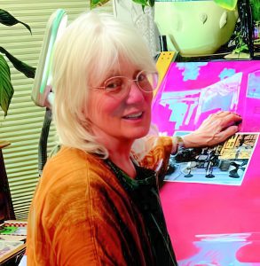 UPPAA An older woman with white hair and glasses sits at a brightly-lit desk, working on a colorful art project. She is smiling and surrounded by plants and various creative supplies.