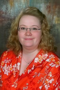 UPPAA A woman with curly blonde hair and glasses is wearing a red floral-patterned blouse. She is smiling softly and posing in front of a brown, textured background.