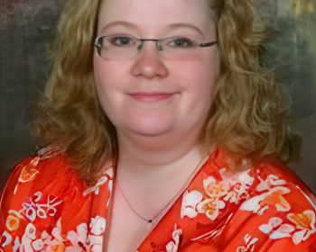 UPPAA A woman with curly blonde hair and glasses is wearing a red floral-patterned blouse. She is smiling softly and posing in front of a brown, textured background.