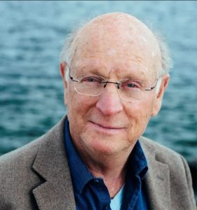UPPAA An older man with glasses, light skin, and thinning hair wears a blue shirt and brown blazer. He stands outdoors in front of a body of water, looking at the camera with a slight smile.