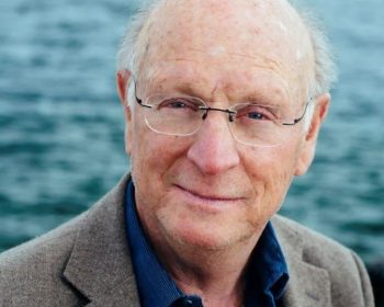 UPPAA An older man with glasses, light skin, and thinning hair wears a blue shirt and brown blazer. He stands outdoors in front of a body of water, looking at the camera with a slight smile.