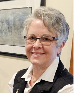 UPPAA An older woman with short gray hair and glasses smiles at the camera. She is wearing a white collared shirt and a black vest. A framed winter landscape painting hangs on the wall behind her.