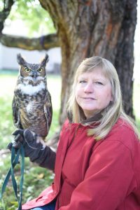 UPPAA A woman with blonde hair wearing a red jacket sits outdoors in front of a tree, holding a great horned owl perched on her gloved hand. The background includes green grass and blurred trees.