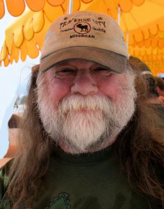 UPPAA An older man with long hair and a white beard smiles at the camera. He is wearing a tan cap that says "Traverse City, Michigan," and stands in front of bright yellow umbrellas.