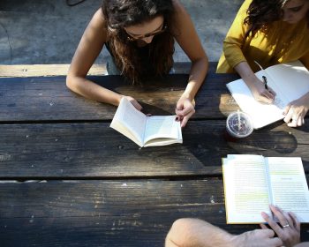 UPPAA Two people sit at a wooden picnic table, one reading a book and the other writing in a notebook. An open book with highlighted text and a plastic cup with a drink are also on the table.
