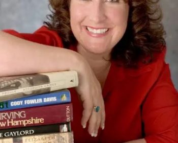 UPPAA A woman with brown hair wearing a red top smiles at the camera, resting her arm on a tall stack of books with various covers and titles visible. The background is neutral and softly lit.