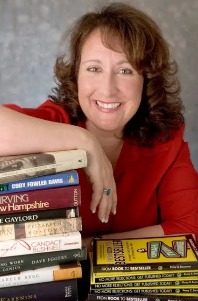 UPPAA A woman with brown hair wearing a red top smiles at the camera, resting her arm on a tall stack of books with various covers and titles visible. The background is neutral and softly lit. UPPAA A woman with brown hair wearing a red top smiles at the camera, resting her arm on a tall stack of books with various covers and titles visible. The background is neutral and softly lit.