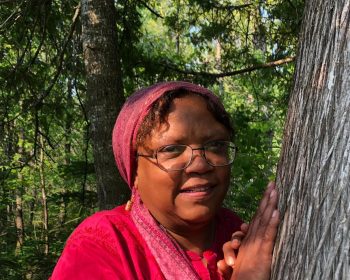 UPPAA A woman wearing glasses, a red headscarf, and a red shirt stands in a forest with her hands resting on the trunk of a tree. Sunlight filters through the trees in the background.