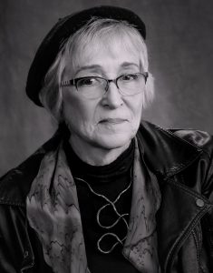 UPPAA An older woman with short light hair wearing glasses, a beret, a patterned scarf, a dark top, and a leather jacket, poses against a plain dark background.