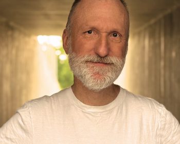 UPPAA A middle-aged man with a white beard and short hair stands in a tunnel, wearing a plain white t-shirt. Soft sunlight is visible in the background, creating a warm glow.