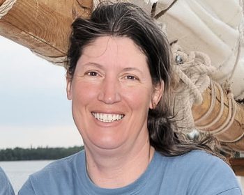 UPPAA A woman with long brown and gray hair, wearing a blue shirt, smiles at the camera in front of a wooden sailboat mast and rigging, with water and trees visible in the background.
