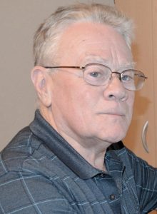UPPAA An older man with short gray hair, glasses, and a striped dark polo shirt looks toward the camera with a neutral expression. A wooden cabinet and beige wall are visible in the background.
