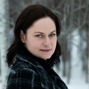 UPPAA A woman with pale skin, dark shoulder-length hair, and blue eyes stands outdoors in a snowy forest, wearing a dark plaid coat and a black turtleneck. The background shows bare trees and snow.