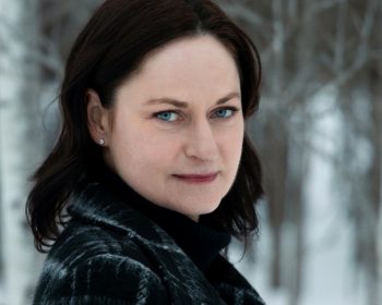 UPPAA A woman with pale skin, dark shoulder-length hair, and blue eyes stands outdoors in a snowy forest, wearing a dark plaid coat and a black turtleneck. The background shows bare trees and snow.