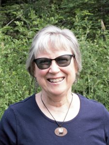 UPPAA An older woman with short gray hair wears black sunglasses, a navy blue shirt, earrings, and a necklace, smiling while standing outdoors in front of leafy green foliage.