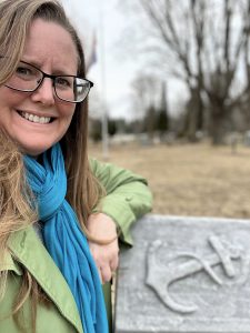 UPPAA A person with long hair, glasses, and a blue scarf smiles at the camera while standing outdoors next to a stone marker with a carved deer antler design. Trees and a cloudy sky are in the background.