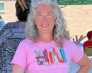 UPPAA A woman with curly gray hair stands outdoors, smiling. She wears a pink T-shirt that shows books on a shelf and the words "I'M WITH THE BANNED." There are people and a tent in the background.