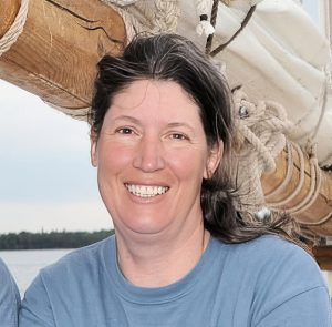 UPPAA A woman with long brown hair and a blue shirt smiles at the camera while standing outdoors near wooden nautical rigging and water.