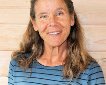 UPPAA A woman with long, wavy brown hair and a blue striped shirt is smiling at the camera. She is standing indoors in front of a light wood-paneled wall.