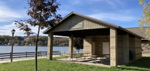 UPPAA A covered pavilion with picnic tables sits on grass near a lake. A small tree and lamppost are in the foreground. There are autumn trees and houses on the far shore under a blue sky with thin clouds.