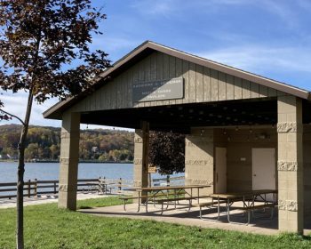 UPPAA A covered pavilion with picnic tables sits on grass near a lake. A small tree and lamppost are in the foreground. There are autumn trees and houses on the far shore under a blue sky with thin clouds.