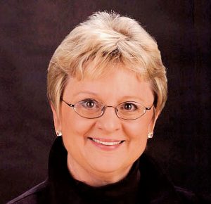 UPPAA A middle-aged person with short blonde hair, glasses, and light skin, wearing a black top and pearl earrings, is smiling in front of a dark background.