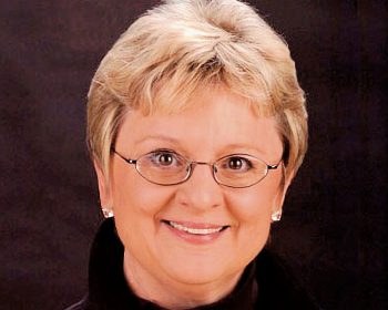 UPPAA A middle-aged person with short blonde hair, glasses, and light skin, wearing a black top and pearl earrings, is smiling in front of a dark background.