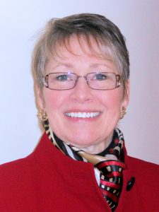 UPPAA A person with short, light hair and glasses smiles at the camera. They are wearing a red jacket, gold earrings, and a patterned scarf with black, white, and red colors. The background is plain and light-colored.