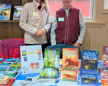 UPPAA Two women stand behind a table displaying books titled “Gift Horse,” “Straw Horse,” and “Moose Willow Mystery,” with a price list sign between them.